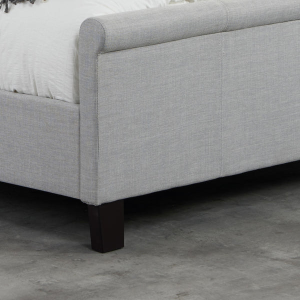 Gray upholstered bed frame with dark wooden legs on a concrete floor.
