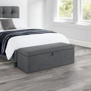 Gray storage bench in front of a bed in a bedroom with wooden flooring.