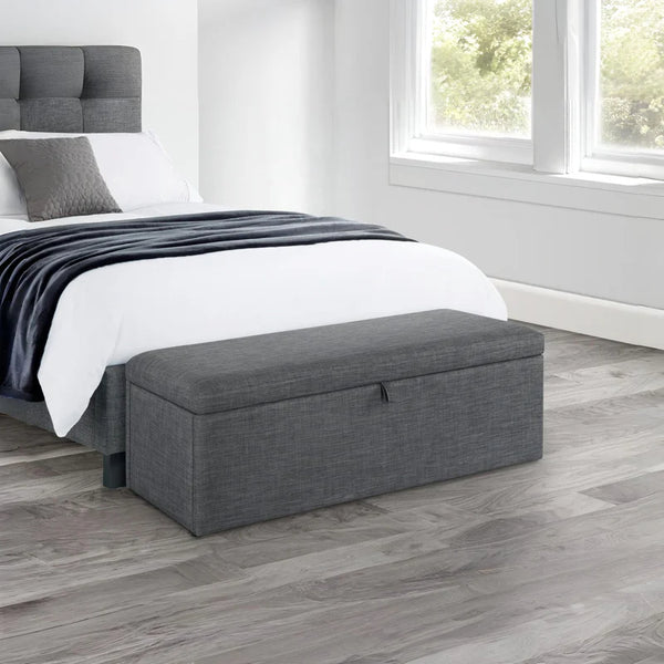 Gray storage bench in front of a bed in a bedroom with wooden flooring.