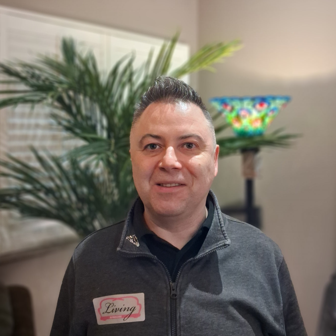 Man wearing a gray jacket with a logo, standing indoors with plants and a colorful lamp in the background.