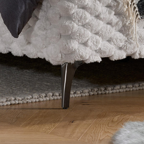 Close-up of a textured white bed with a dark wooden leg and gray carpeted area.