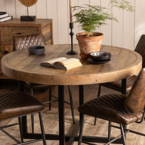 Round wooden dining table with four brown chairs in a rustic setting.