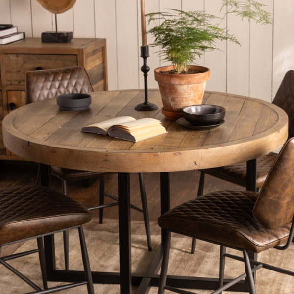 Round wooden dining table with four brown chairs in a rustic setting.